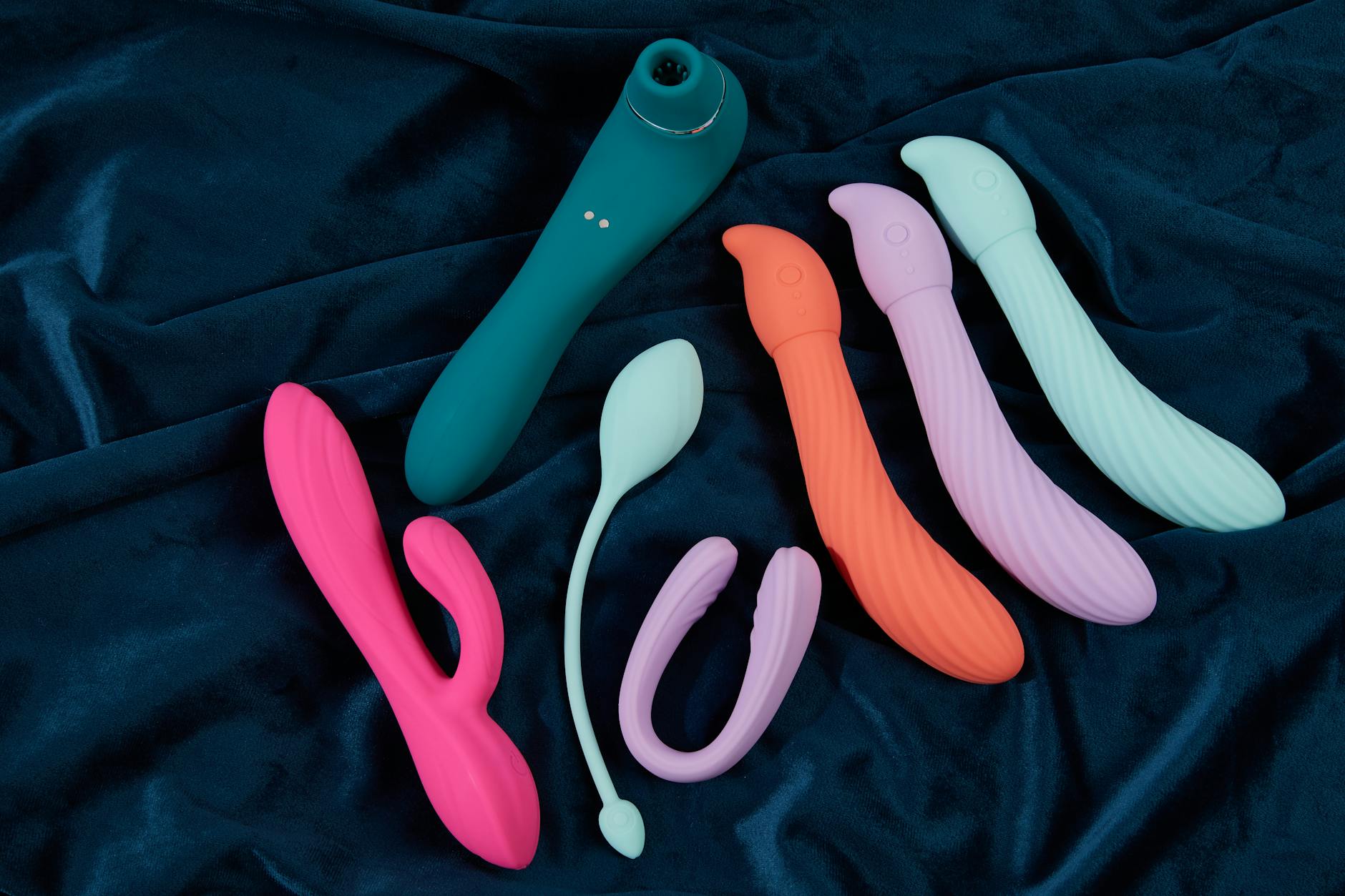 A close-up of a vibrant collection of silicone sex toys on dark blue fabric, showcasing various colors and shapes