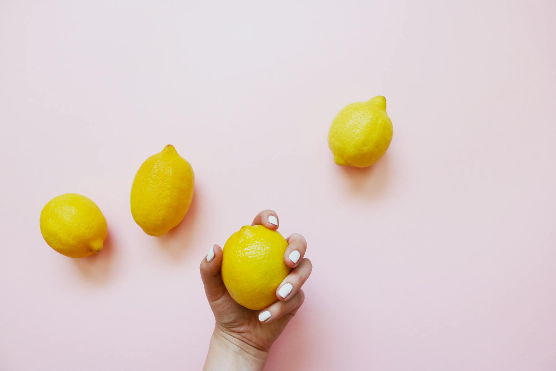 A hand holding a fresh lemon surrounded by additional lemons on a soft pink background