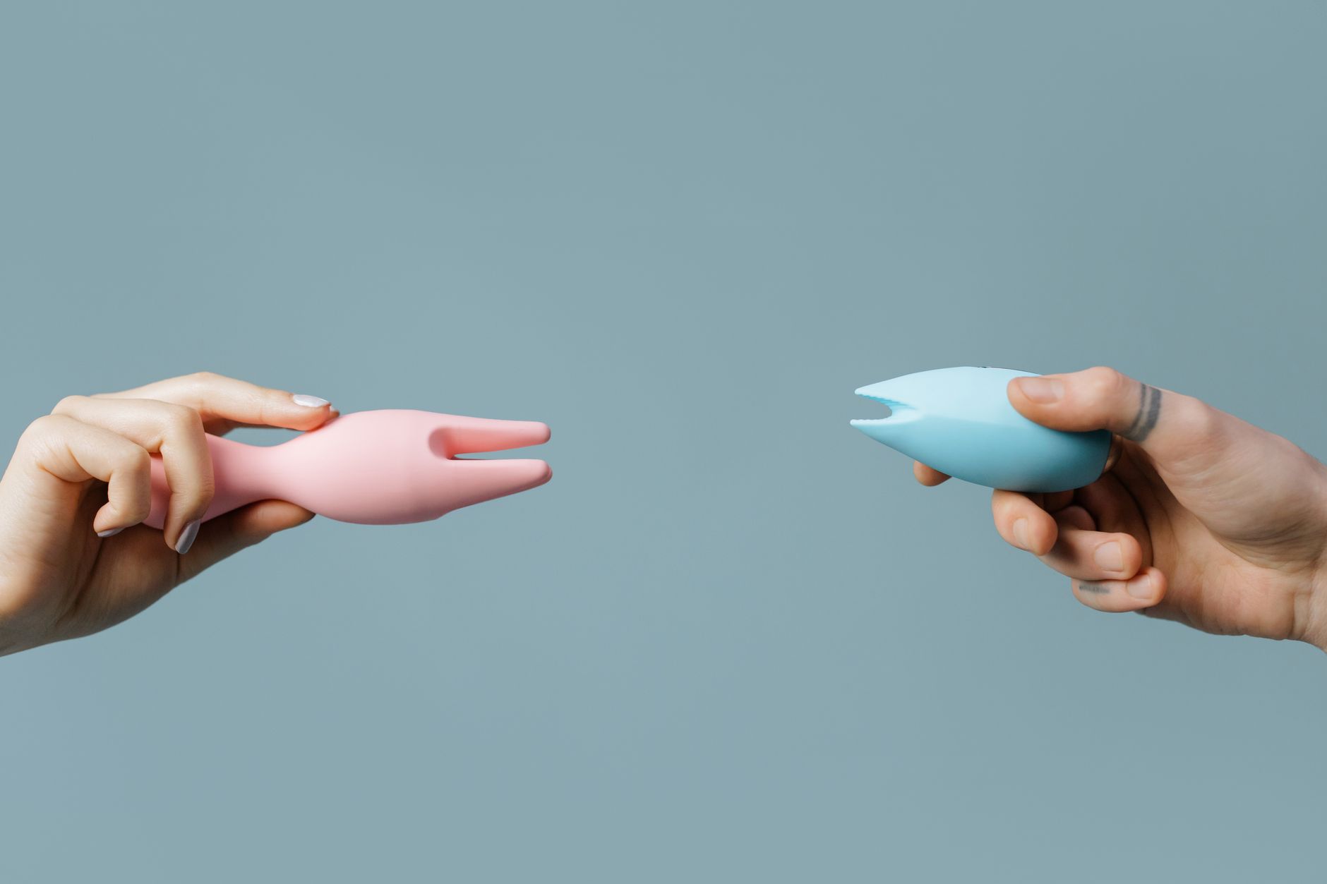 Two hands holding colorful silicone vibrators together against a soft pastel background.