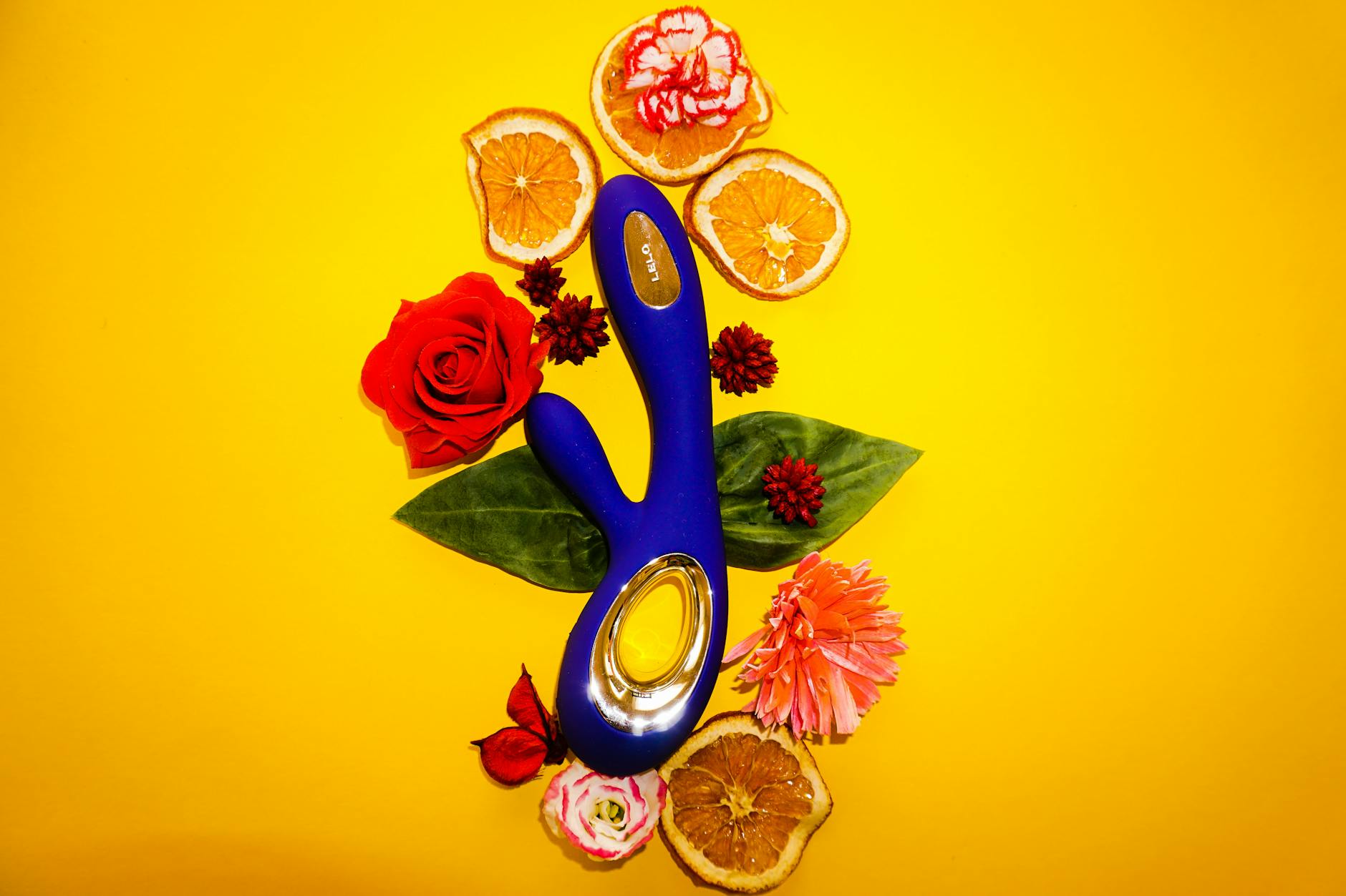 Colorful clitoral vibrator surrounded by flowers and citrus on a bright yellow background